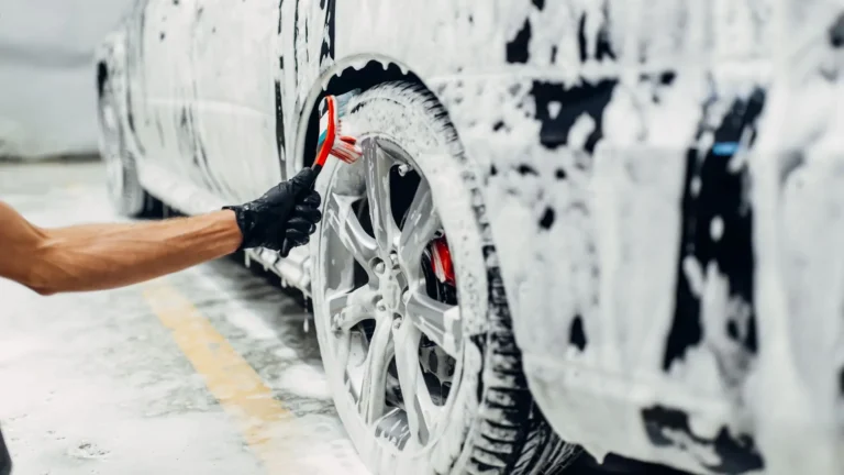 Person reinigt einen Reifen eines schwarzen Autos von Hand mit einem Schwamm bei Autowäsche